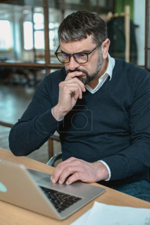 Téléchargez les photos : Thoughtful male entrepreneur working in office, doing web research while looking at laptop screen - en image libre de droit