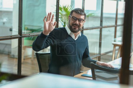 Téléchargez les photos : Portrait of smiling man in glasses sitting at his desk in the office while waving to his colleague - en image libre de droit