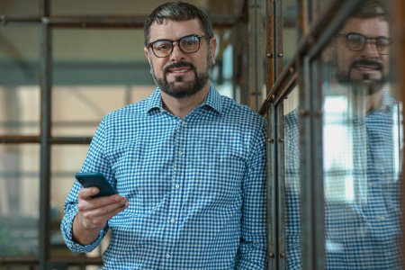 Téléchargez les photos : Portrait de gérant masculin heureux habillé chemise à carreaux et tenant smartphone à l'intérieur - en image libre de droit