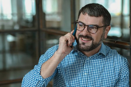 Téléchargez les photos : Positive Caucasian man in casual shirt talking by the smartphone in the modern office, copy space - en image libre de droit