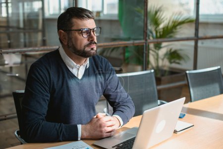 Téléchargez les photos : Homme adulte concentré dans des lunettes assis sur le lieu de travail et utilisant un ordinateur portable - en image libre de droit
