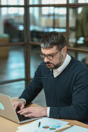 Téléchargez les photos : Occupé homme d'affaires caucasien dans des lunettes en utilisant son ordinateur portable tout en tapant sur le lieu de travail - en image libre de droit