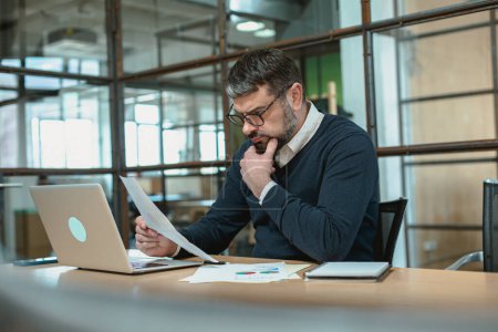 Téléchargez les photos : Homme adulte dans des lunettes assis sur le lieu de travail tout en travaillant avec des papiers dans le bureau - en image libre de droit