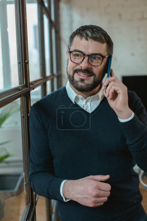 Foto de Hombre adulto guapo hablando en el teléfono móvil y sonriendo mientras está de pie en la oficina - Imagen libre de derechos