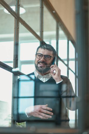 Téléchargez les photos : Sourire homme caucasien dans des lunettes profiter de la conversation sur le lieu de travail tout en utilisant le téléphone - en image libre de droit
