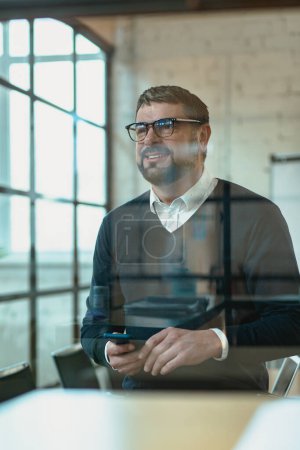 Téléchargez les photos : Homme adulte heureux dans des lunettes tenant téléphone portable pendant le travail dans le bureau - en image libre de droit