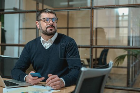 Téléchargez les photos : Homme caucasien dans des lunettes de travail avec ordinateur portable et documents tout en tenant smartphone - en image libre de droit
