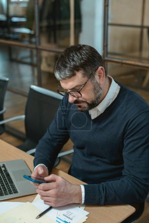Téléchargez les photos : Homme occupé dans des lunettes en utilisant un smartphone et en tapant un message alors qu'il était assis dans le bureau - en image libre de droit