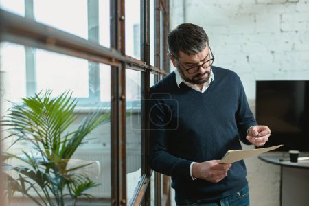 Téléchargez les photos : Homme d'affaires à lunettes tenant des documents alors qu'il se tenait debout dans le bureau - en image libre de droit