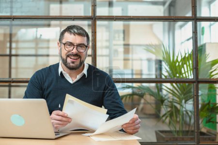 Téléchargez les photos : Homme d'affaires souriant dans des lunettes travaillant sur un ordinateur portable et tenant des papiers tout en étant assis à une table en bois - en image libre de droit