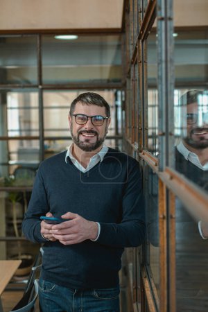Téléchargez les photos : Homme d'affaires adulte heureux utilisant un smartphone dans un bureau tout en posant sur la caméra - en image libre de droit