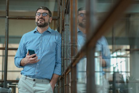 Téléchargez les photos : Portrait d'un homme heureux avec des lunettes debout dans un bureau lumineux et tenant un téléphone portable - en image libre de droit
