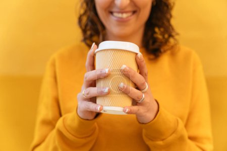 Photo for Crop unrecognizable positive female in sweater with paper cup of takeaway hot drink in hands standing on yellow background on street - Royalty Free Image