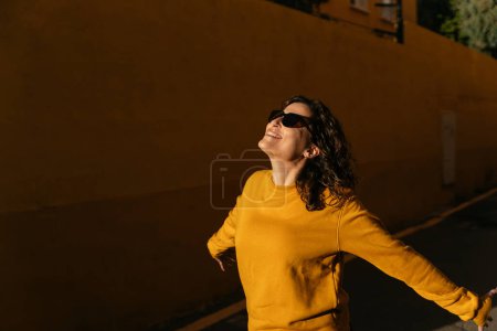 Foto de Mujer alegre con el pelo oscuro en suéter amarillo y gafas de sol paseando por la calle soleada con los brazos extendidos cerca de la pared en la ciudad - Imagen libre de derechos