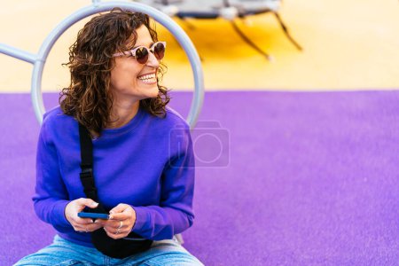 Téléchargez les photos : Femme positive avec des cheveux bouclés dans des vêtements décontractés et des lunettes de soleil messagerie texte sur téléphone portable moderne tout en étant assis sur une aire de jeux en ville - en image libre de droit