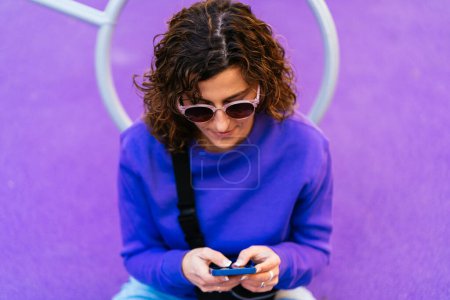 Photo for From above of concentrated female with curly hair and sunglasses in casual wear text messaging on cellphone while sitting on playground - Royalty Free Image
