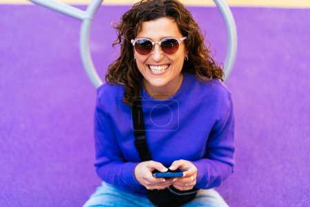 Téléchargez les photos : Femme gaie aux cheveux bouclés foncés dans des lunettes de soleil messagerie texte sur téléphone portable tout en étant assis sur une aire de jeux colorée sur la rue de la ville - en image libre de droit