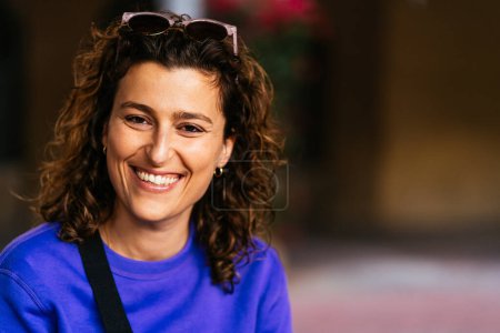 Foto de Mujer optimista con gafas de sol sobre cabello rizado oscuro en suéter casual mirando a la cámara con sonrisa mientras está sentada sobre un fondo borroso - Imagen libre de derechos