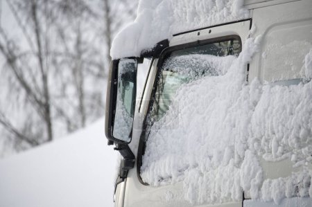 Foto de Primer plano de un parabrisas de camión blanco, capucha, y espejo lateral están notablemente cubiertos de nieve espesa y esponjosa, el vehículo está parcialmente enterrado bajo la nieve - Imagen libre de derechos
