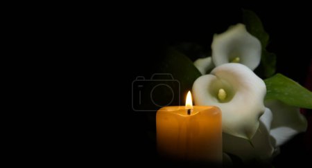 Téléchargez les photos : Bougie à cire brûlante unique irradiant une douce flamme à côté de la fleur de lys de calla blanche dans un environnement sombre. Vue rapprochée avec espace de copie gratuit - en image libre de droit
