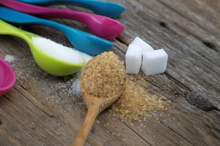 Brown cane sugar and white sugar in colorful plastic measuring spoons spilled around on a rustic wooden surface, measuring utensils in a kitchen setting