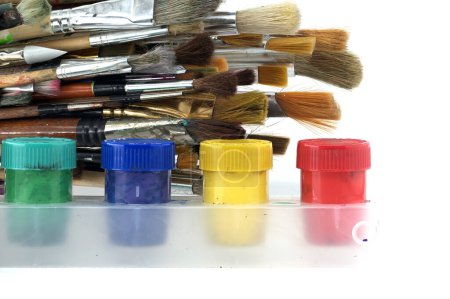 Array of paintbrushes of different sizes and colors alongside cans of acrylic paint with visible colors including red, yellow, blue, and green arranged on a white surface