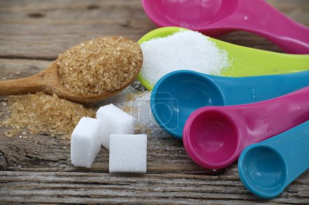 Brown cane sugar and white sugar in colorful plastic measuring spoons spilled around on a rustic wooden surface, measuring utensils in a kitchen setting