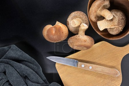 Bowl containing fresh shiitake mushrooms, wooden cutting board with knife on it and dark towel arranged on black background