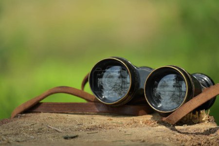 Pair of black binoculars with a brown leather strap is resting atop a large brown tree stump, lenses of the binoculars reflecting the image of trees, background is blurred with dominant green hues