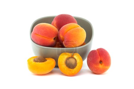 Bowl filled with fresh whole apricots and one cut in half to reveal its interior, isolated on a white background