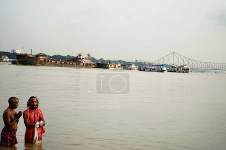 Téléchargez les photos : Deux personnes prient pour Dieu dans le ghat ganga de Kolkata montrant le pont Howrah à Bakcground pendant mahalaya Tarpan - en image libre de droit