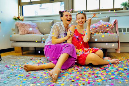 Photo for Two friends having party at home sitting on floor holding party blowers with confetti falling around them. LGBT, non-binary - Royalty Free Image