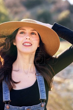 Photo for Smiling country girl wearing bib overalls holding straw hat on head - Royalty Free Image