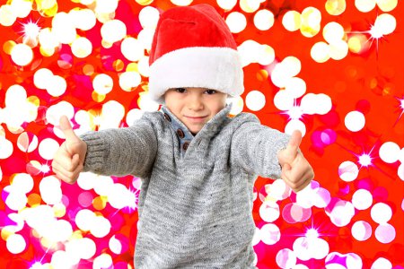 Foto de Divertido niño de cinco años con un sombrero de Santa para la fiesta de Navidad con dos pulgares hacia arriba. Fondo bokeh colorido festivo. - Imagen libre de derechos