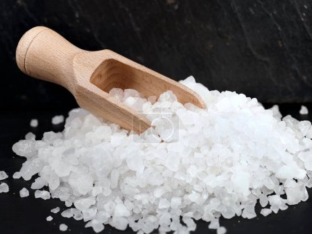 Photo for Heap of coarse sea salt with wooden spice scoop on black slate background, close up of natural salt crystals for cooking. - Royalty Free Image