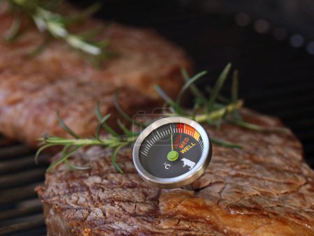 delicious beef steak on grill grate with a meat thermometer showing doneness of rare, medium and well done, close up of grilling a perfect steak on barbecue.
