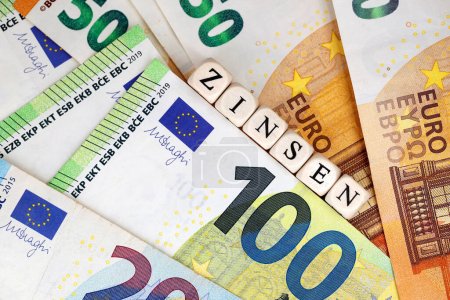 Vue du dessus du mot allemand pour les taux d'intérêt écrits avec des cubes de lettres en bois sur divers billets en euros, image conceptuelle pour le financement ou l'investissement avec les intérêts associés.