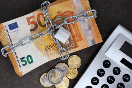 A stack of cash with 50 euro banknotes is locked with a chain and padlock next to a calculator and coins, concept for the safe investment of fixed-term deposits or financial security in the future.