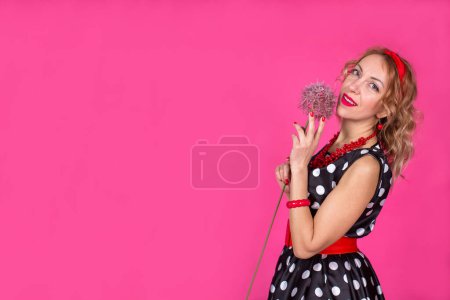 Téléchargez les photos : Une fille blonde dans une robe noire à pois blancs avec des accessoires rouges se tient sur un fond rose avec une énorme belle fleur d'allium. Pin-up. - en image libre de droit