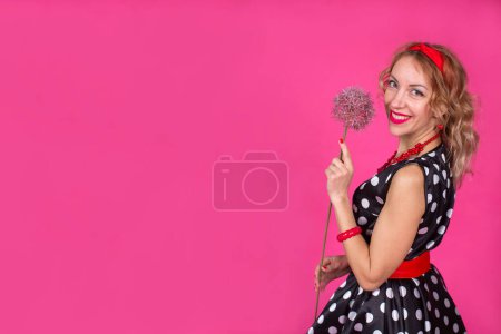 Téléchargez les photos : Une fille blonde dans une robe noire à pois blancs avec des accessoires rouges se tient sur un fond rose avec une énorme belle fleur d'allium. Pin-up. - en image libre de droit