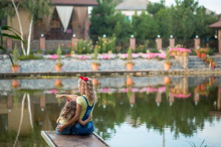 Téléchargez les photos : Mère blonde et fille de trois ans en short en denim avec des bretelles à nœuds rouges s'assoient sur une jetée sur la rivière au coucher du soleil - en image libre de droit