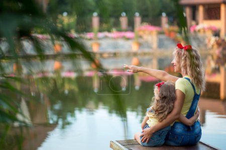 Blonde mother and three-year-old daughter in denim shorts with straps with red bows sit hugging on a pier on the river at sunset