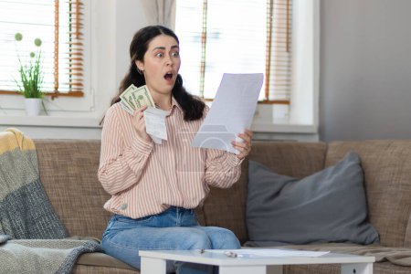 Téléchargez les photos : Purchase of real estate. Shocked young caucasian woman sitting on sofa, holds money and reading documents. Concept of leasing, business and mortgage. - en image libre de droit