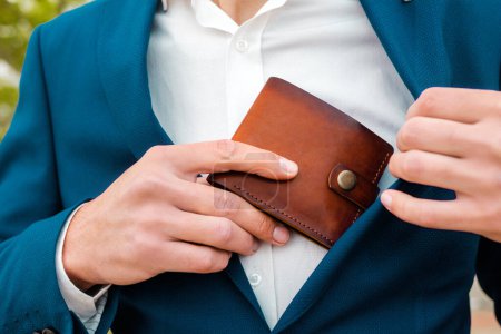 Photo for A businessman in a blue jacket and white shirt take off a brown leather wallet in a pocket. Close-up of the hands. The concept of style and fashion. - Royalty Free Image