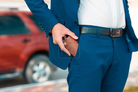 A businessman in a blue suit get a leather key bag from the pants. Defocused car on the background. Close-up of the hands. The concept of style and fashion.