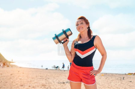 Téléchargez les photos : Sports et entraînement. Bonne femme adulte en tenue de sport, posant sur la plage avec un haltère dans les mains. Ciel et mer en arrière-plan. Espace de copie. - en image libre de droit