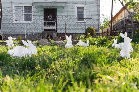 Photo for The concept of Easter holidays. Decorative Handicrafts handmade in the form of white rabbits and colorful eggs lying on the grass. House in the background. Copy space. - Royalty Free Image