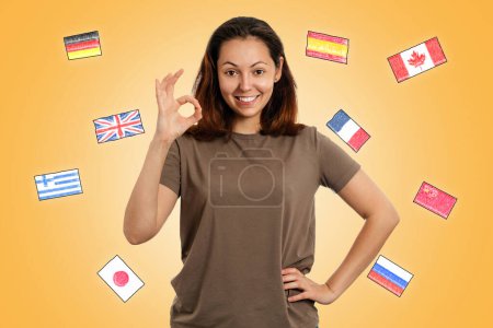 Tag der englischen Sprache. Eine junge lächelnde Frau macht eine OK-Geste. Gelber Hintergrund mit Flaggen verschiedener Länder. Das Konzept des Erlernens von Fremdsprachen.