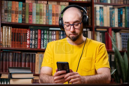 Photo for Audiobooks. A bald man with glasses and headphones uses a smartphone. Shelves with books in the background. The concept of fake news, distance learning, and digital entertainment. - Royalty Free Image