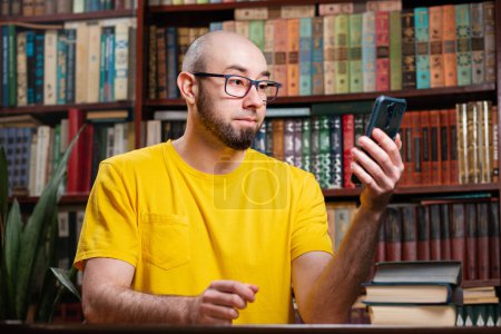 Photo for Communication. A bald bearded man with glasses uses a smartphone. In the background is a library and shelves of books. Concept of social networks, distance learning and quarantine. - Royalty Free Image